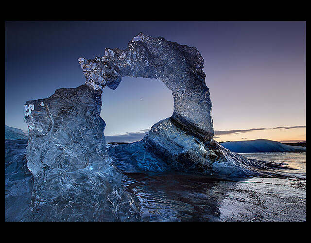 On the frozen Jökulsárlón glacier lake in Iceland, the waxing moon peeks at dusk through a tall iceberg arch with stripes of black volcanic ash