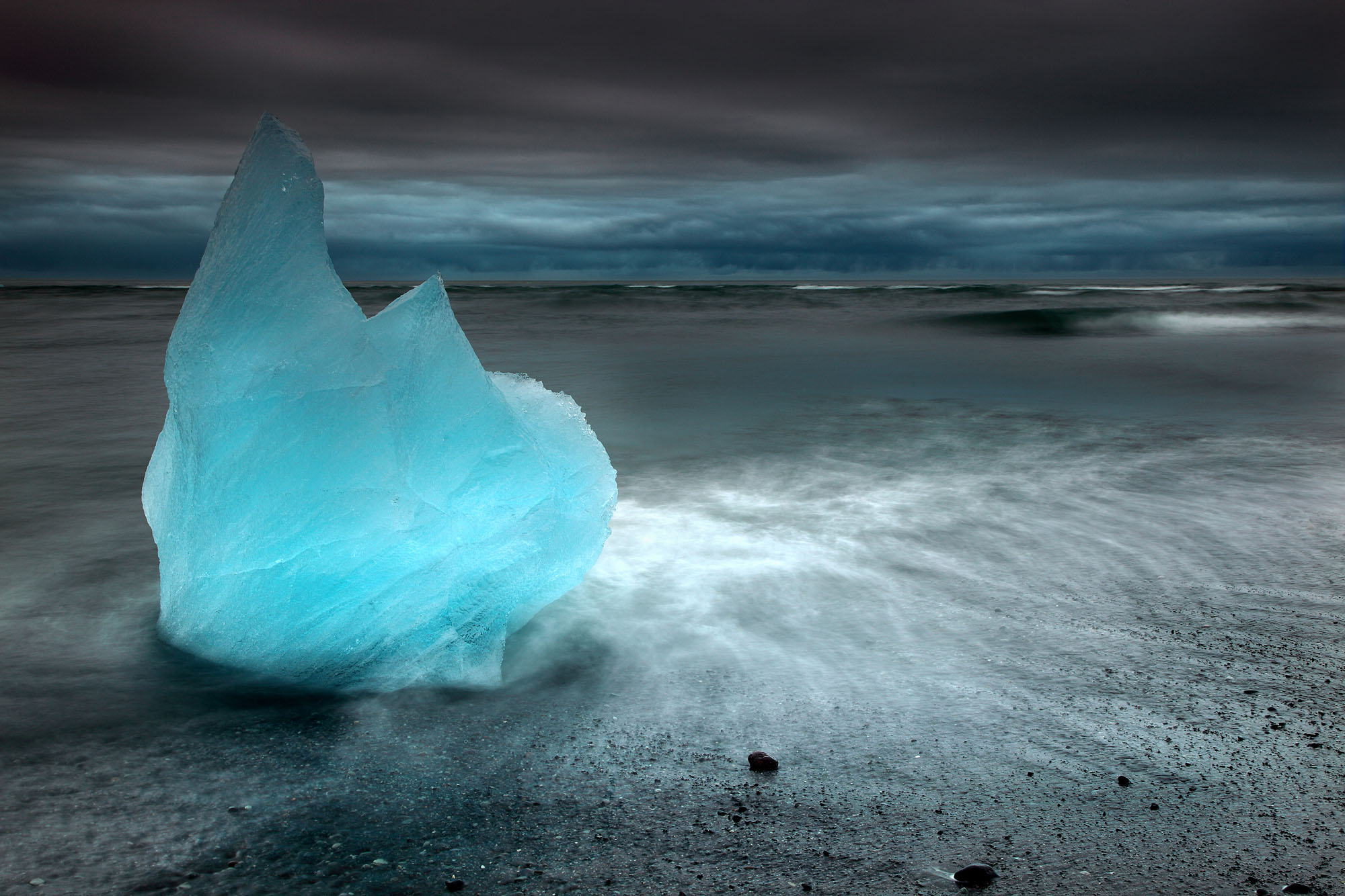 A deep-blue beached iceberg on Jökulsárlón Strandur in Iceland surrounded by waves at low tide close to midnight