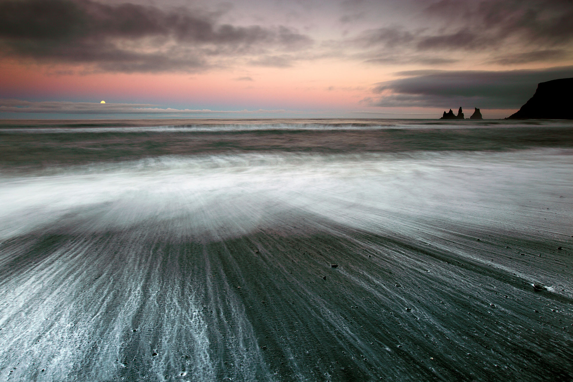 The full moon rising above Vik Strandur beach and the Reynisdrangar rock spires on Iceland at dusk during midsummer while waves create beautiful patterns along the shoreline